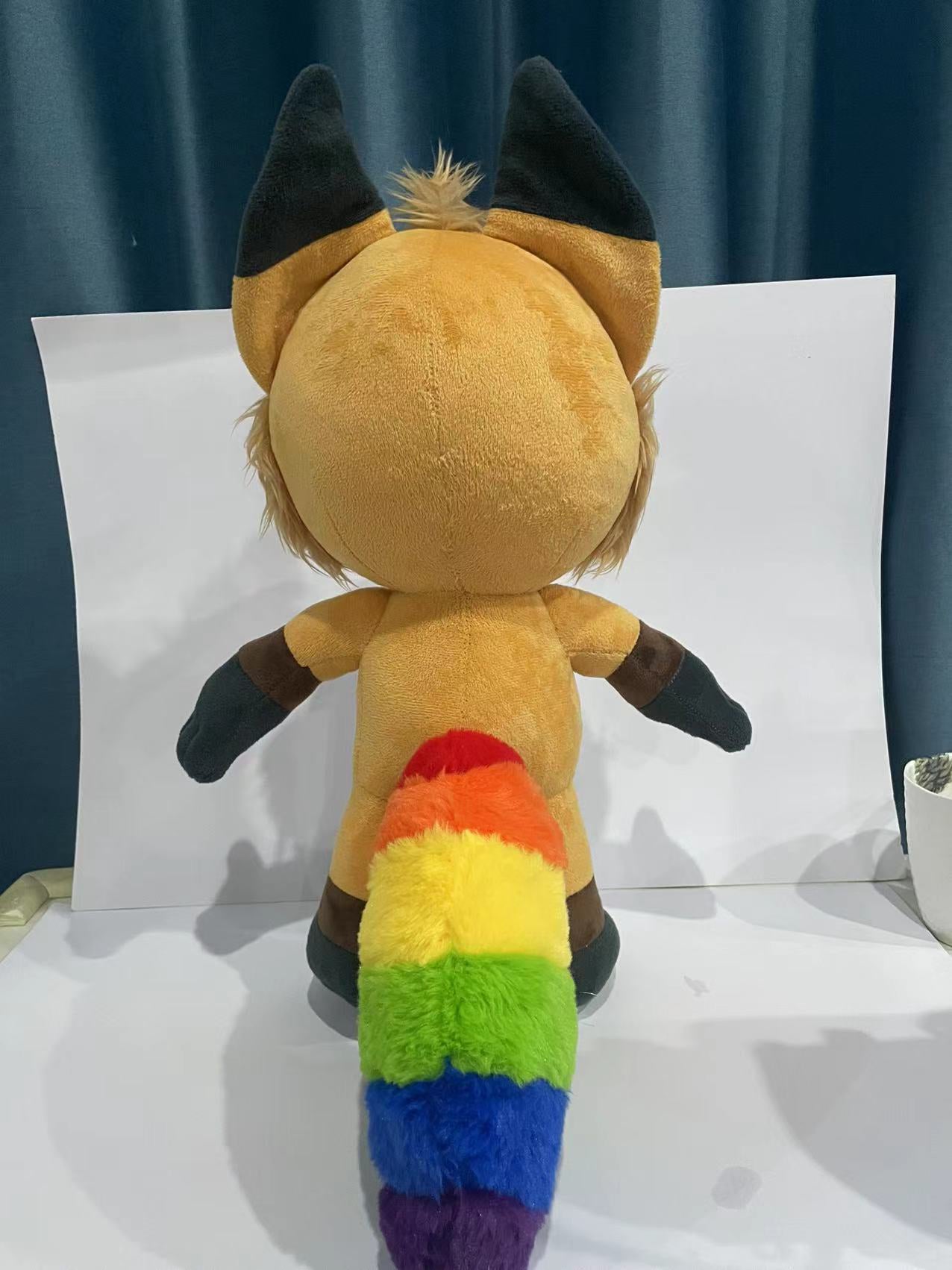 Back view of a plush fox with orange fur with accent colors patterned after the inclusive pride flag